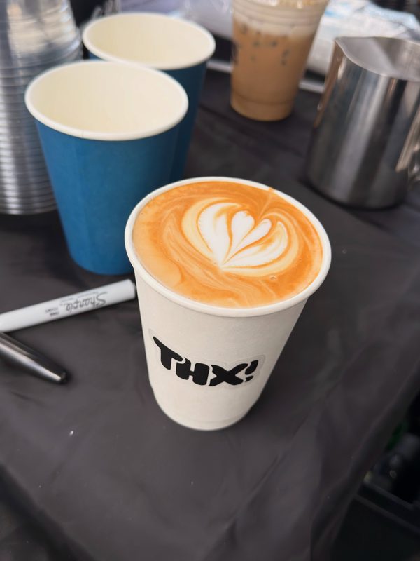 Hot latte with tulip latte art in a thx! cup beside blue cups and a milk pitcher