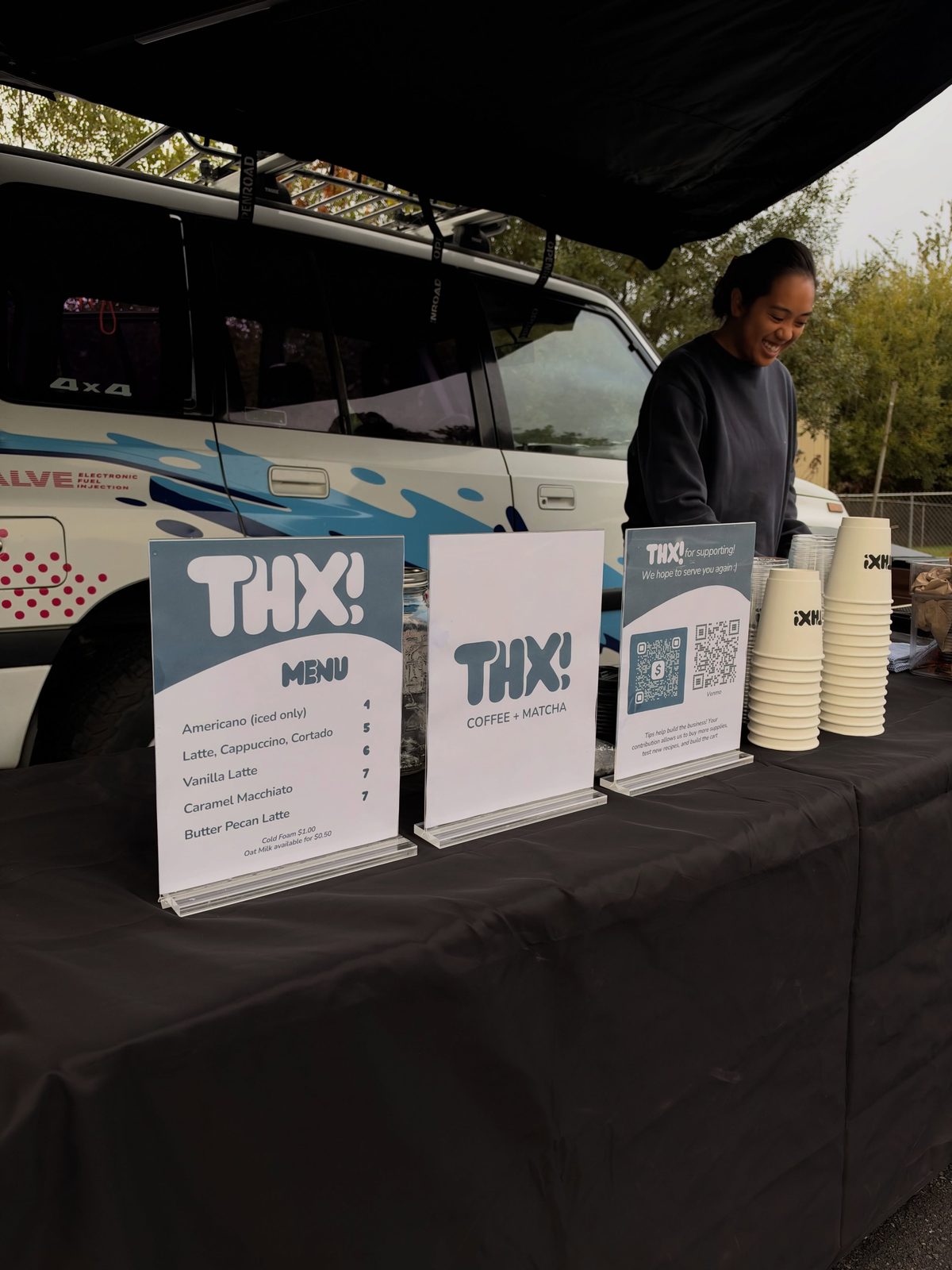 Darlene smiling behind the thx! table with teal-and-white menu stands and stacks of cups